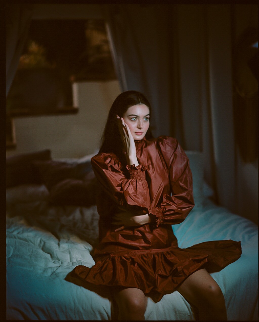 A woman in a red dress sits on a bed with her hand on her cheek.