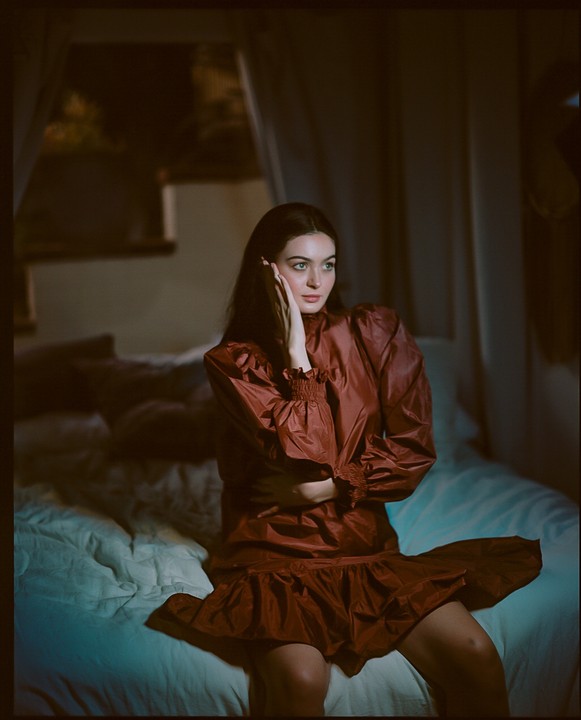 A woman in a red dress sits on a bed with her hand on her cheek.