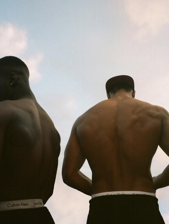Two men are seen from behind, shirtless, against a bright sky.