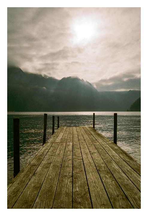 A wooden pier extends into a lake with mountains and a bright sky in the background.