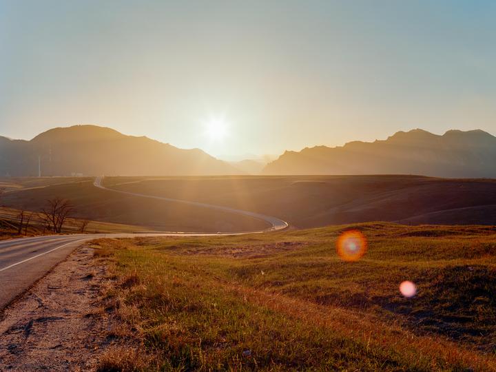 A winding road leads through a grassy landscape towards mountains under a bright sunset.