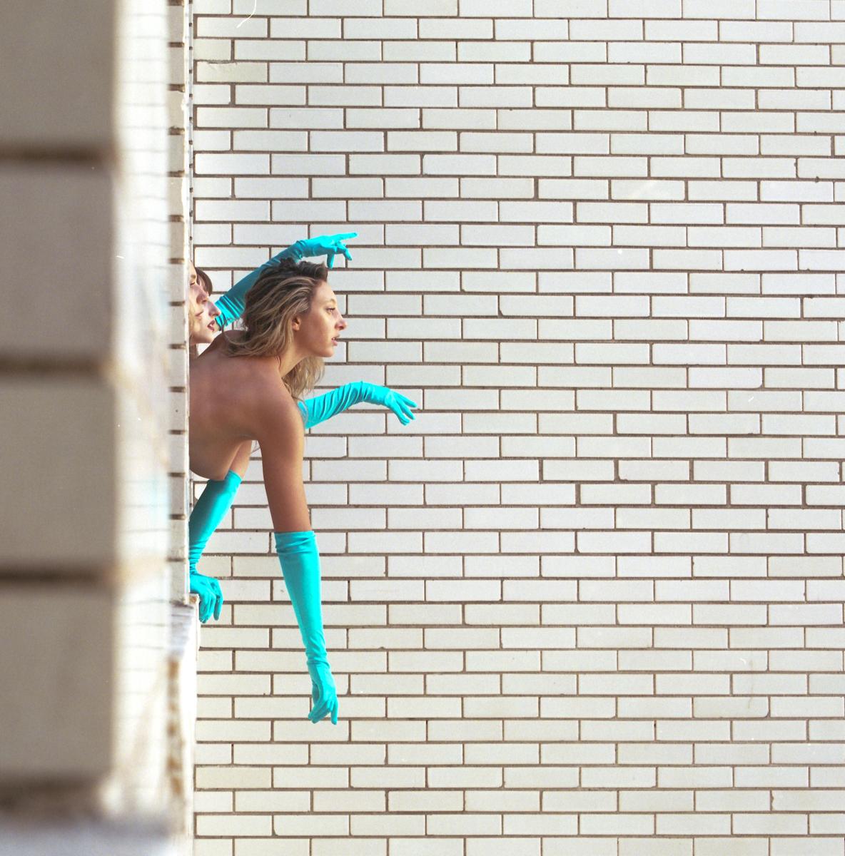 A woman wearing long blue gloves and a matching bottom leans out of a window against a white brick wall.
