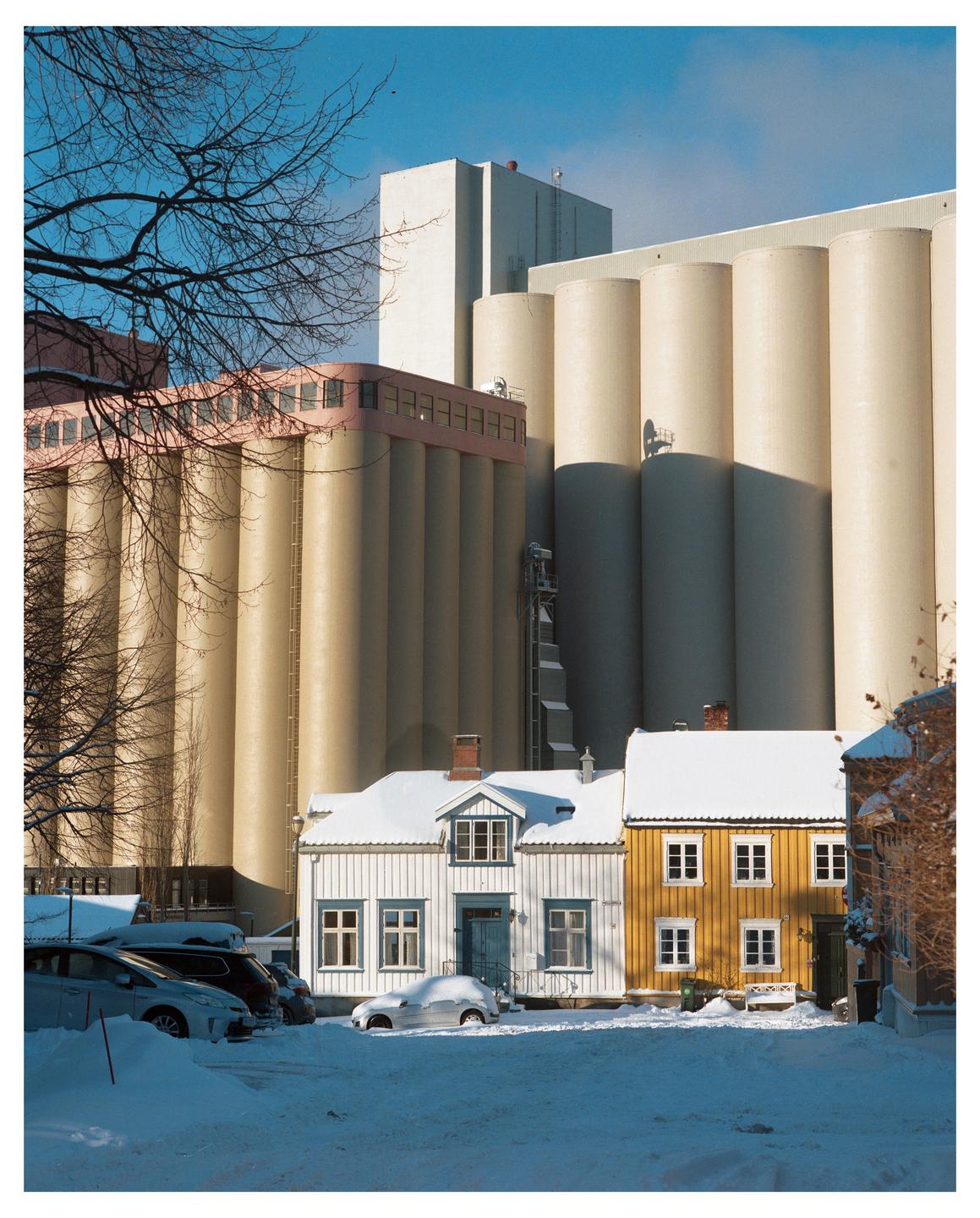 Two snow-covered houses stand in front of large industrial silos under a clear blue sky.
