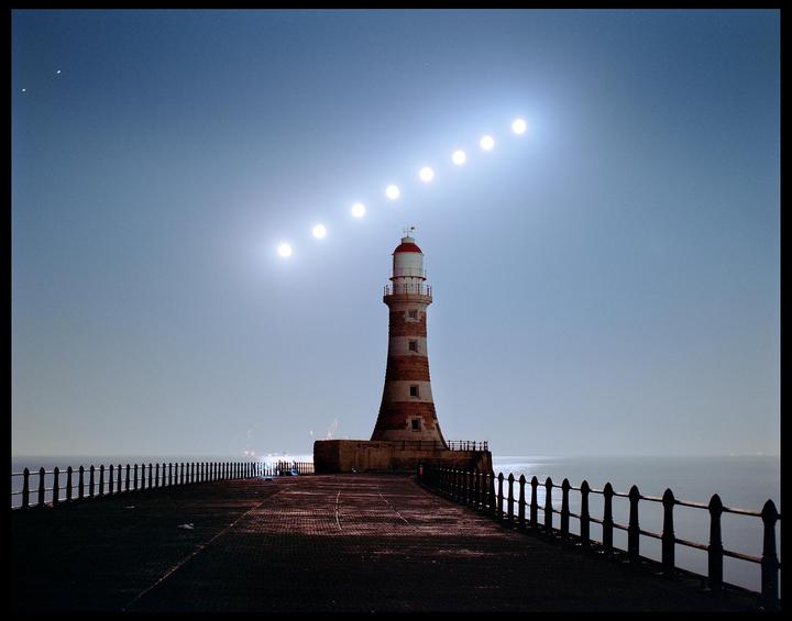 A lighthouse stands on a pier with a sequence of moons rising in the sky.