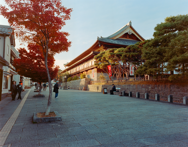 A street with a large temple building and trees with red autumn foliage.
