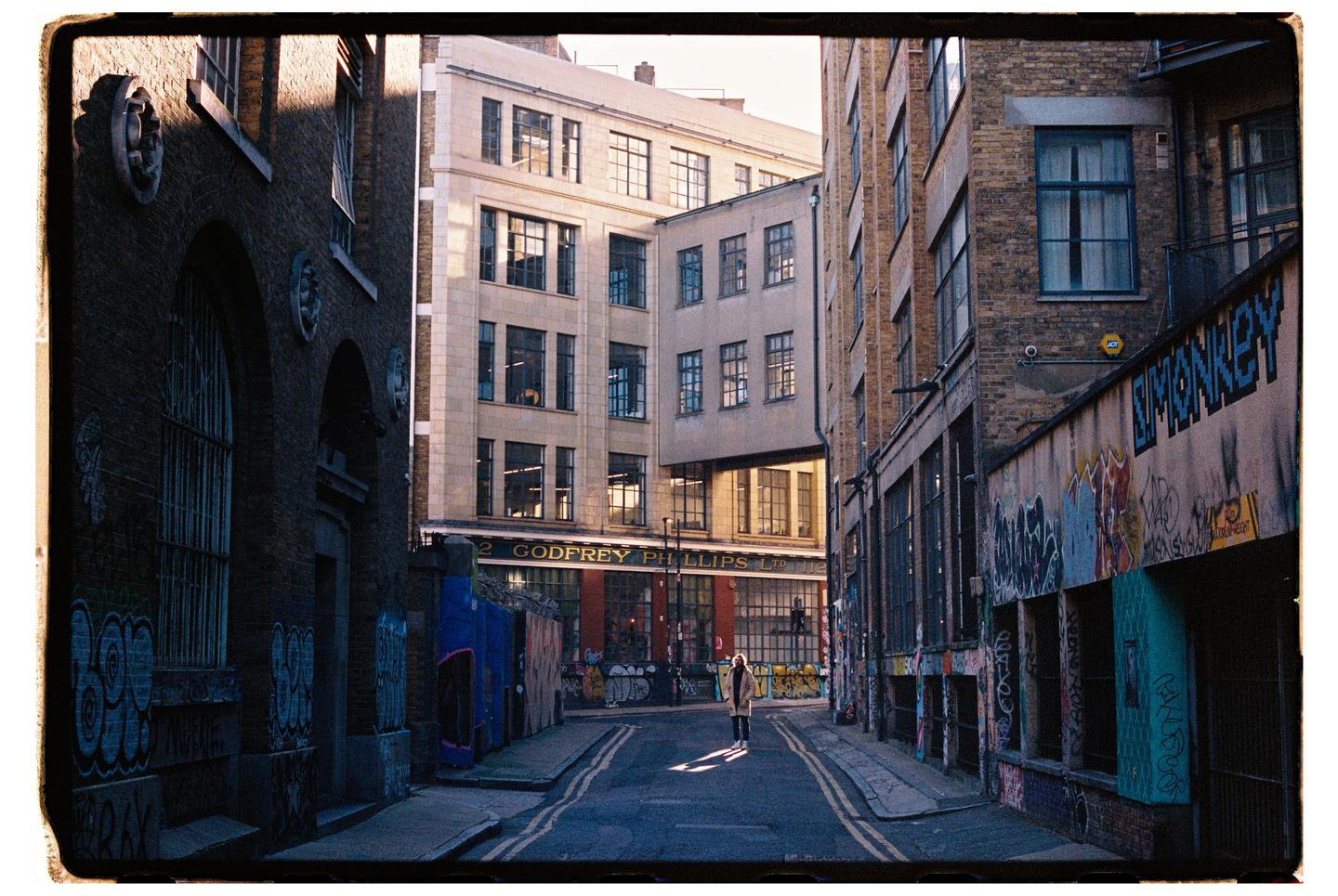 A person stands in a sunlit patch on a narrow street lined with brick buildings and graffiti.