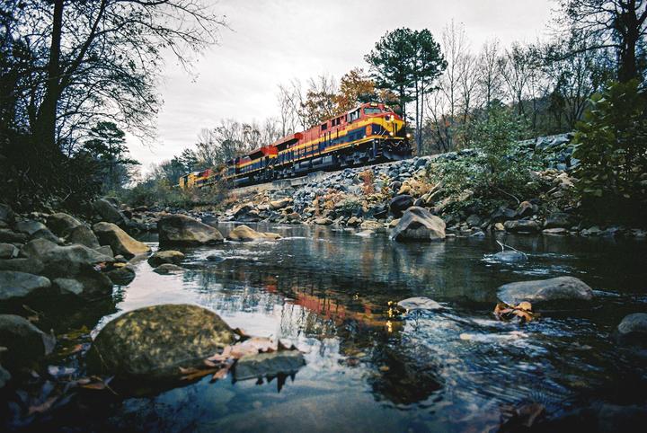 A train travels along a rocky embankment next to a river with autumn foliage on the banks.