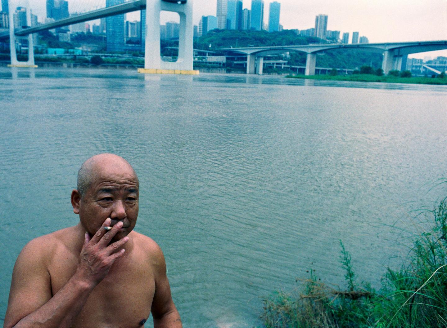 A man smokes a cigarette by a river with bridges and a city in the background.