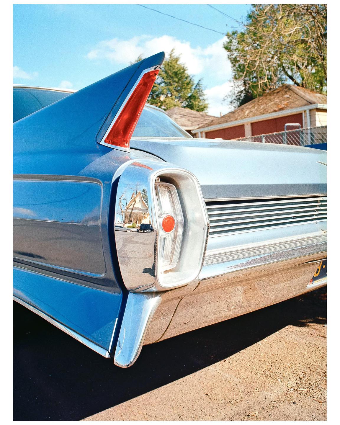 The rear of a blue vintage car with a prominent tail fin and chrome bumper is parked outdoors.