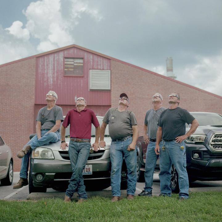 Five men wearing protective glasses look up at the sky in front of a red brick building.