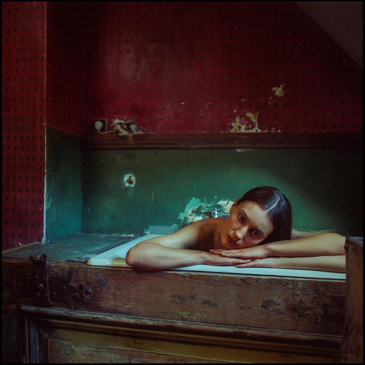 A woman with freckles rests her head on her arms in a bathtub with peeling red and green walls.