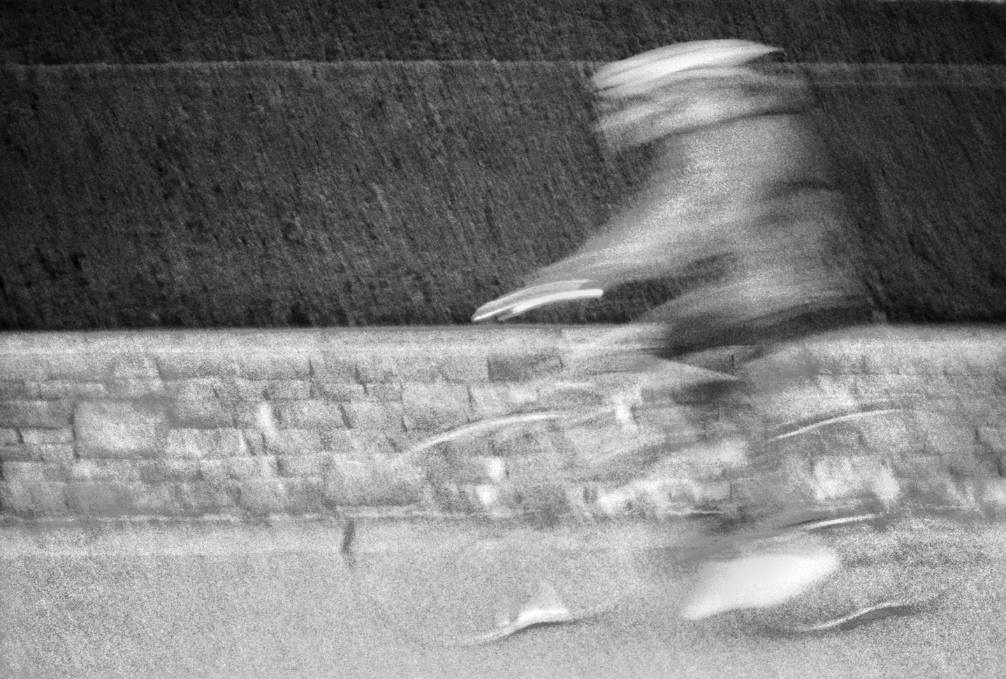 A blurry figure on a bicycle rides past a stone wall and a dark textured surface.