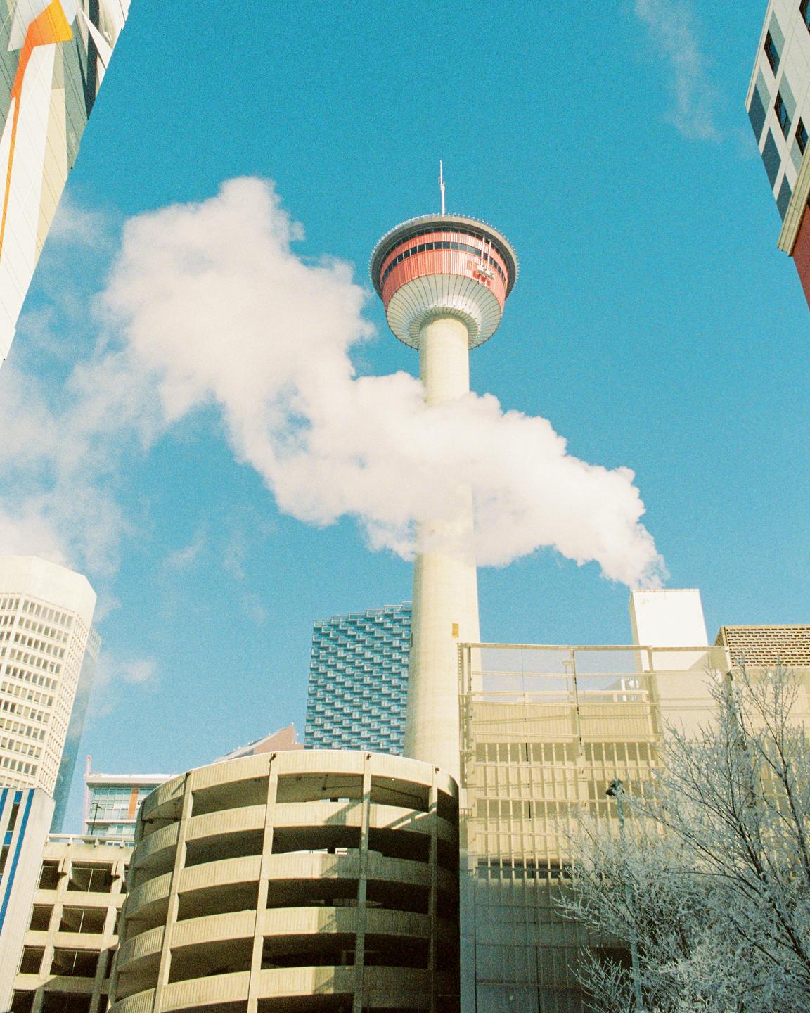 A tall tower with a red observation deck rises above other buildings, with a cloud of smoke or steam partially obscuring its base.