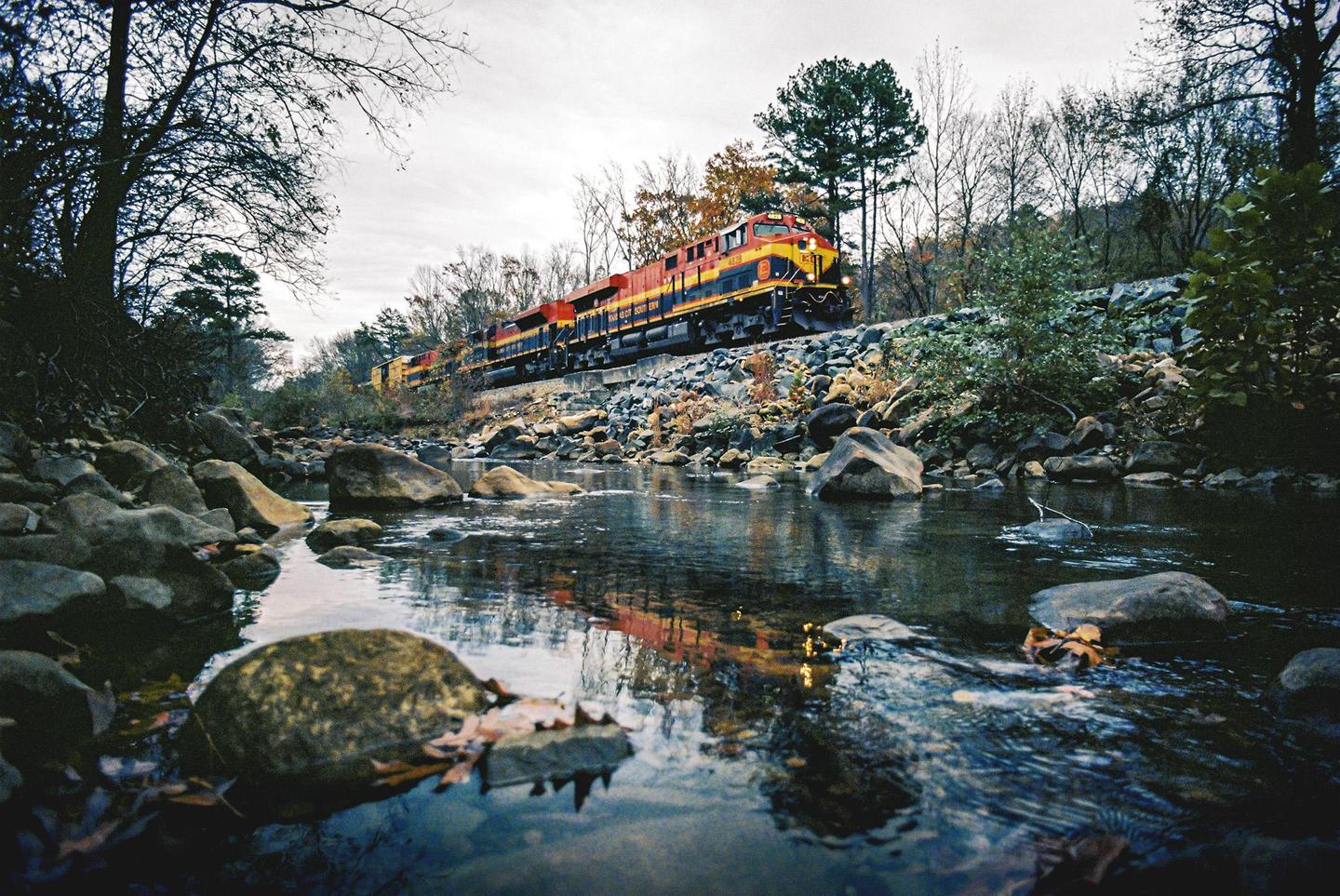 A train travels along a rocky embankment next to a river with autumn foliage on the banks.