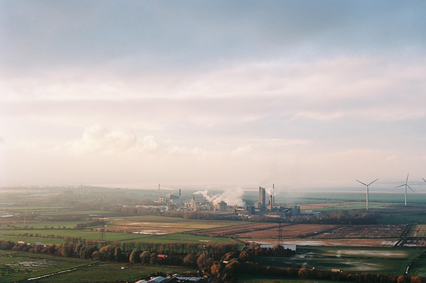 A wide landscape shows an industrial plant with smoking chimneys and wind turbines in a rural area under a hazy sky.