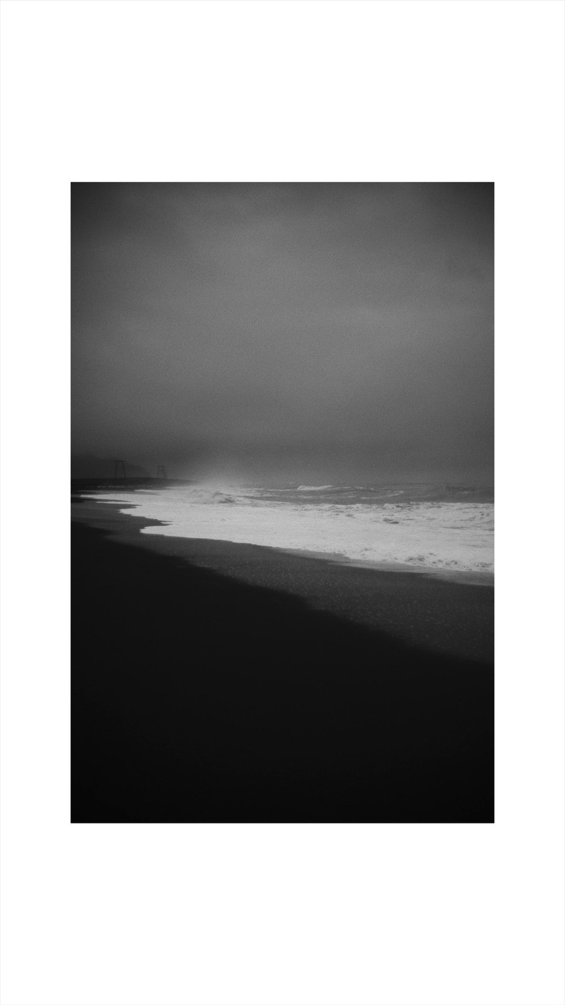 Waves crash on a dark beach under a hazy sky.