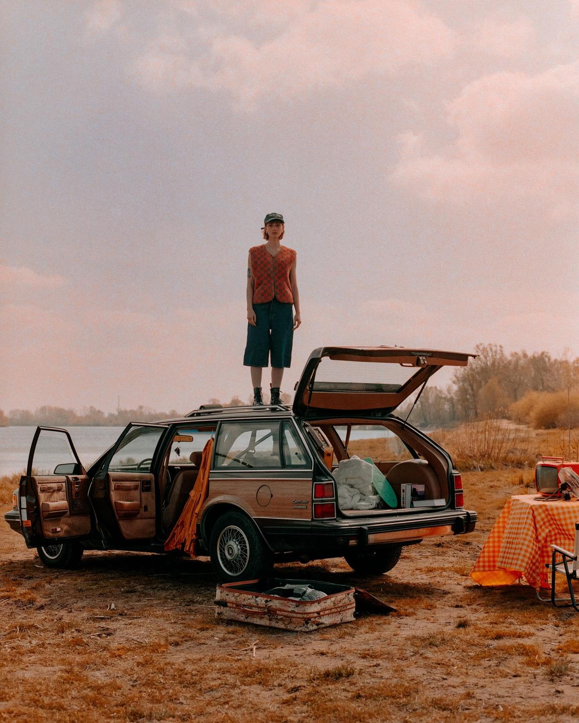 A woman stands on the roof of a station wagon parked by a body of water, with a picnic table and open suitcase nearby.