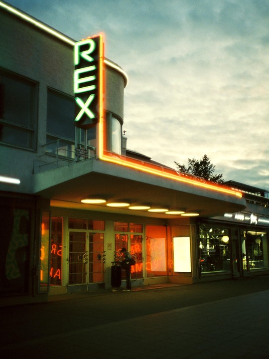 A person stands outside a building with a neon "REX" sign and orange neon lights.