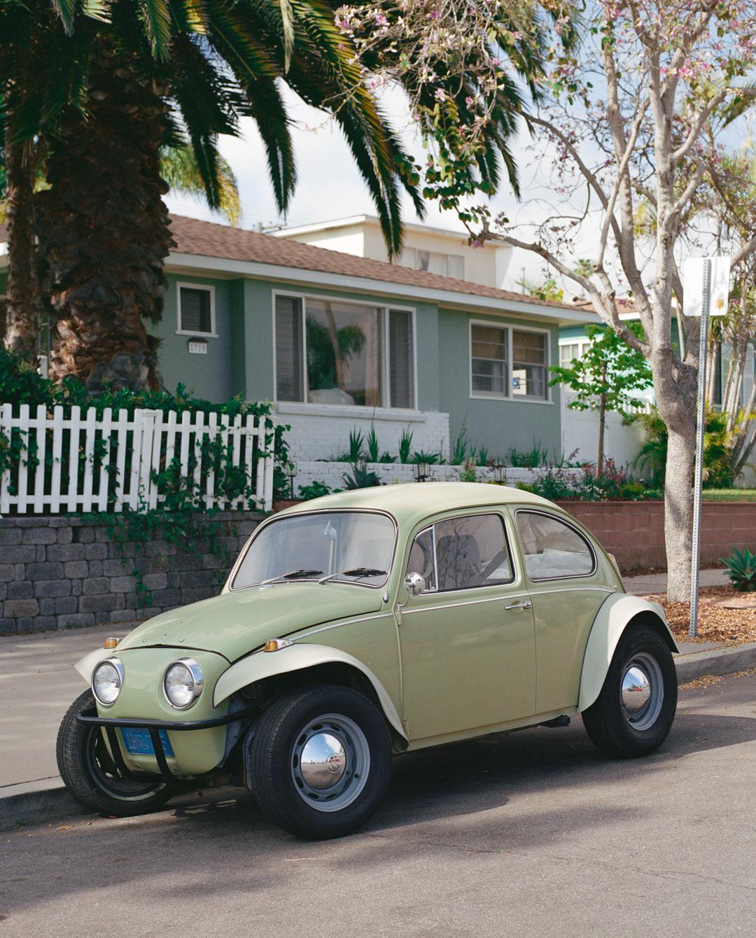 A light green Volkswagen Beetle is parked on a residential street in front of a house with a white picket fence and palm trees.