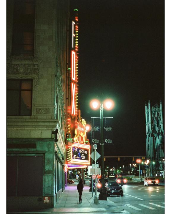 A person walks along a city sidewalk at night next to a building with a large neon sign.
