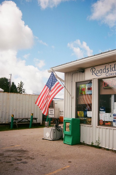 An American flag hangs from the side of a white building with a "Roadside Convenience" sign and an "Open" sign in the window.