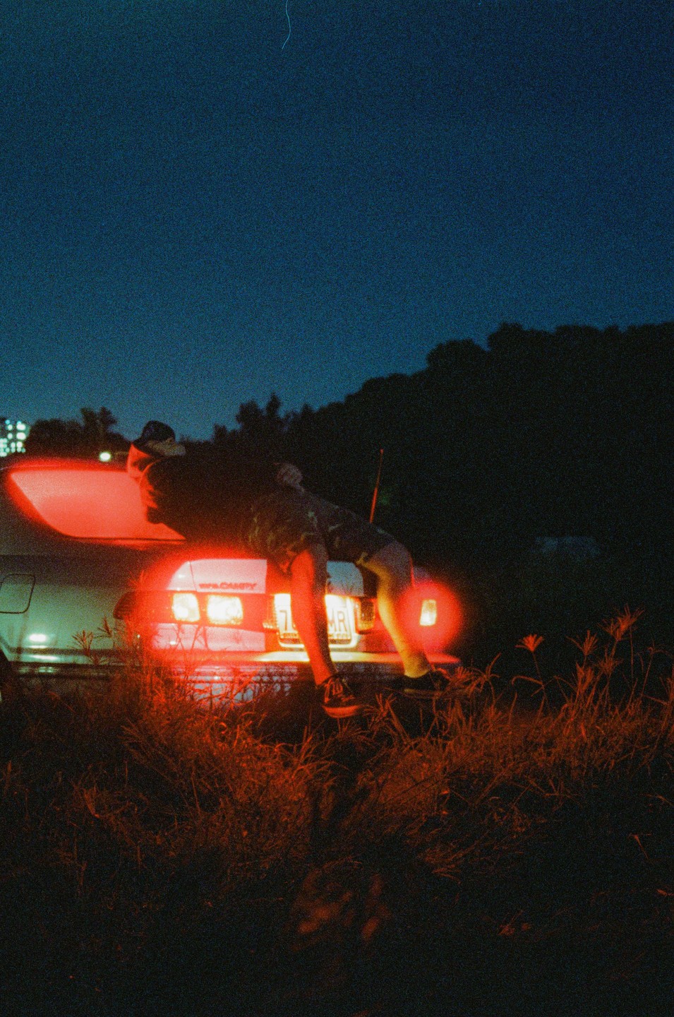 A man leans on the trunk of a car with its red taillights illuminated at night.