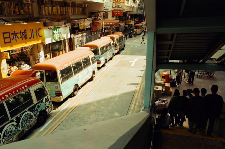 A line of buses is parked on a street next to buildings with storefronts, while people stand on a shaded staircase in the foreground.