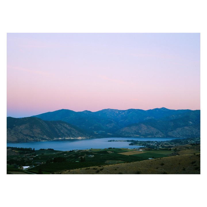 A wide shot of a lake surrounded by mountains and green fields under a lilac sky.