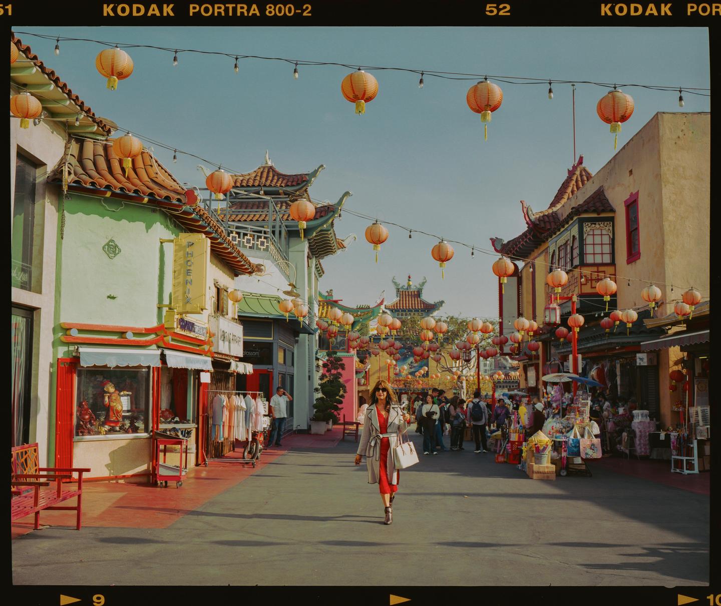 A woman in a red dress and trench coat walks down a street lined with shops and hanging lanterns.