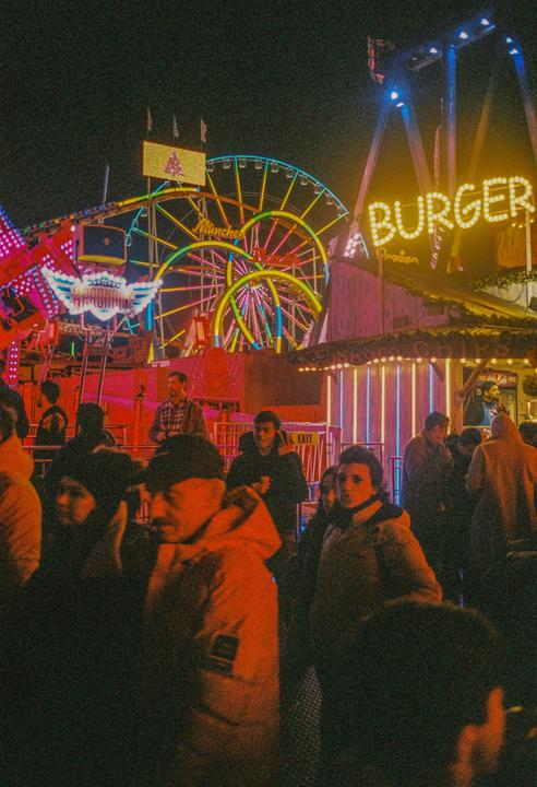A crowd of people stands in front of a brightly lit Ferris wheel and a "BURGER" sign at night.