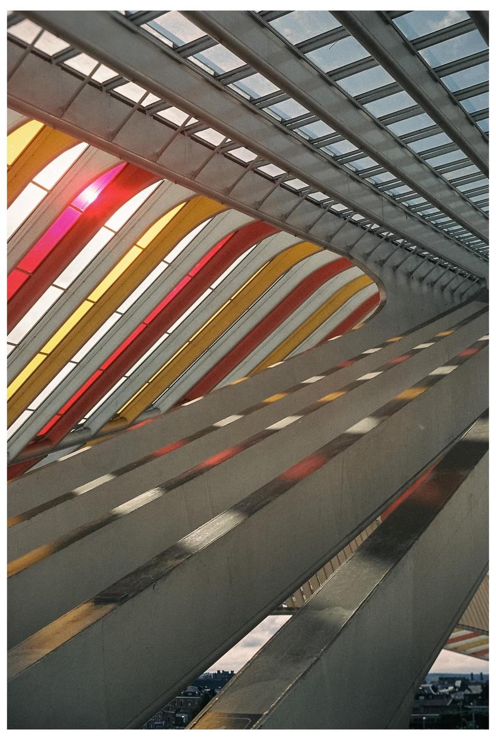 A modern train station features a glass roof with colorful red and yellow panels and strong diagonal lines.