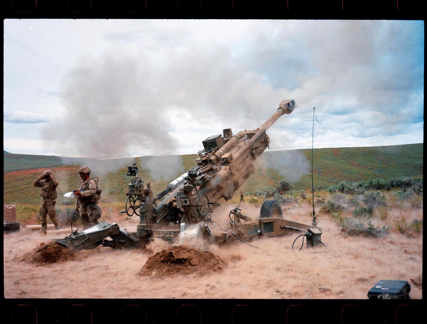 Two men in military uniform stand near a large howitzer cannon that has just fired, creating a cloud of dust and smoke in a desert landscape.