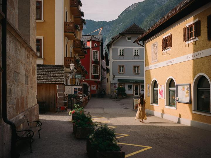 A woman in a long dress walks down a narrow street lined with colorful buildings and mountains in the background.