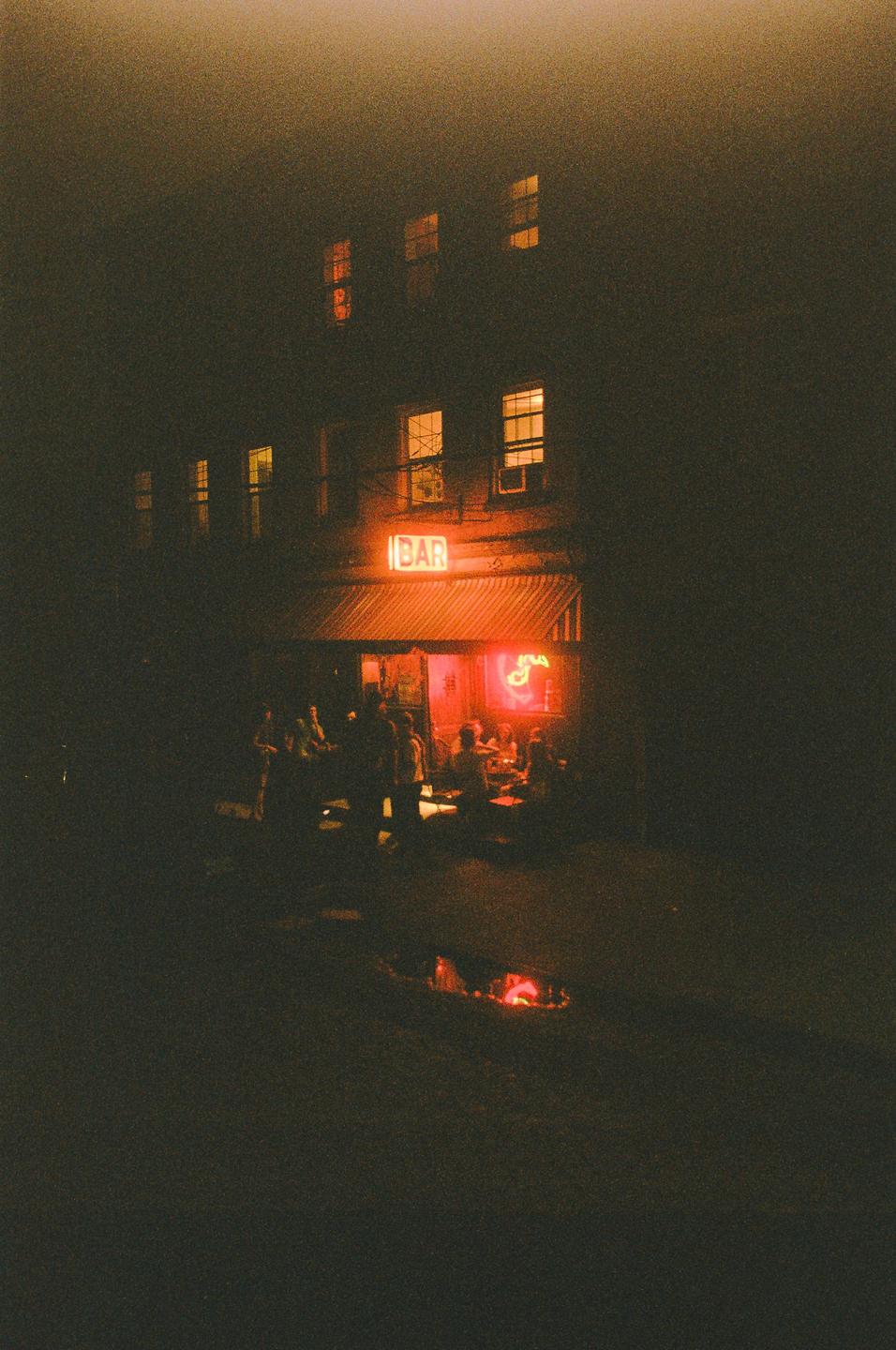 A group of people are gathered outside a bar with a neon sign at night.