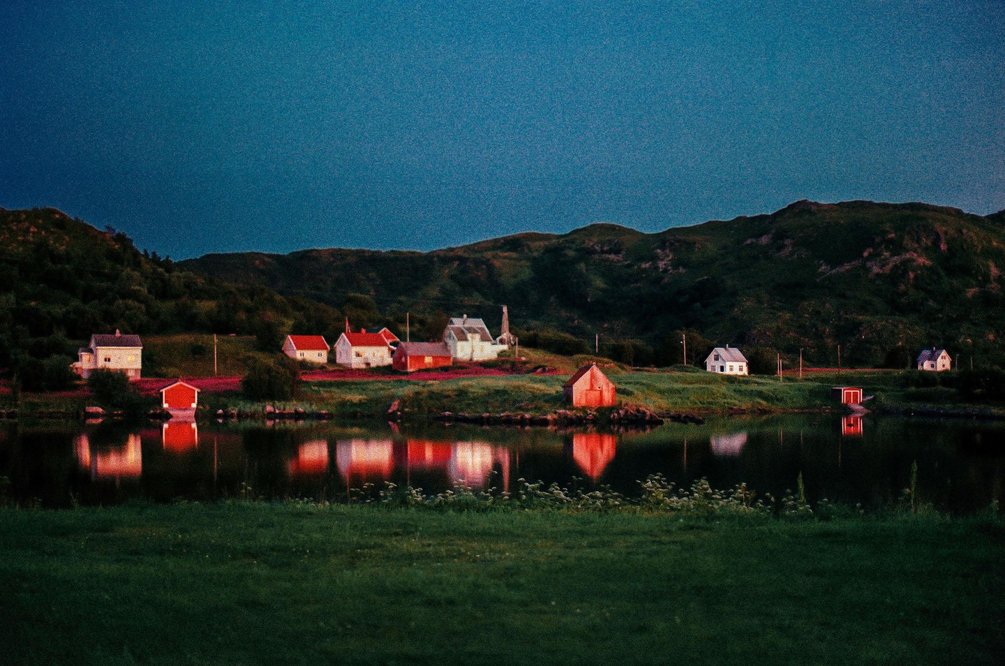 A small village with houses and sheds is reflected in a body of water at dusk, with hills in the background.