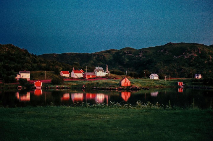 A small village with houses and sheds is reflected in a body of water at dusk, with hills in the background.