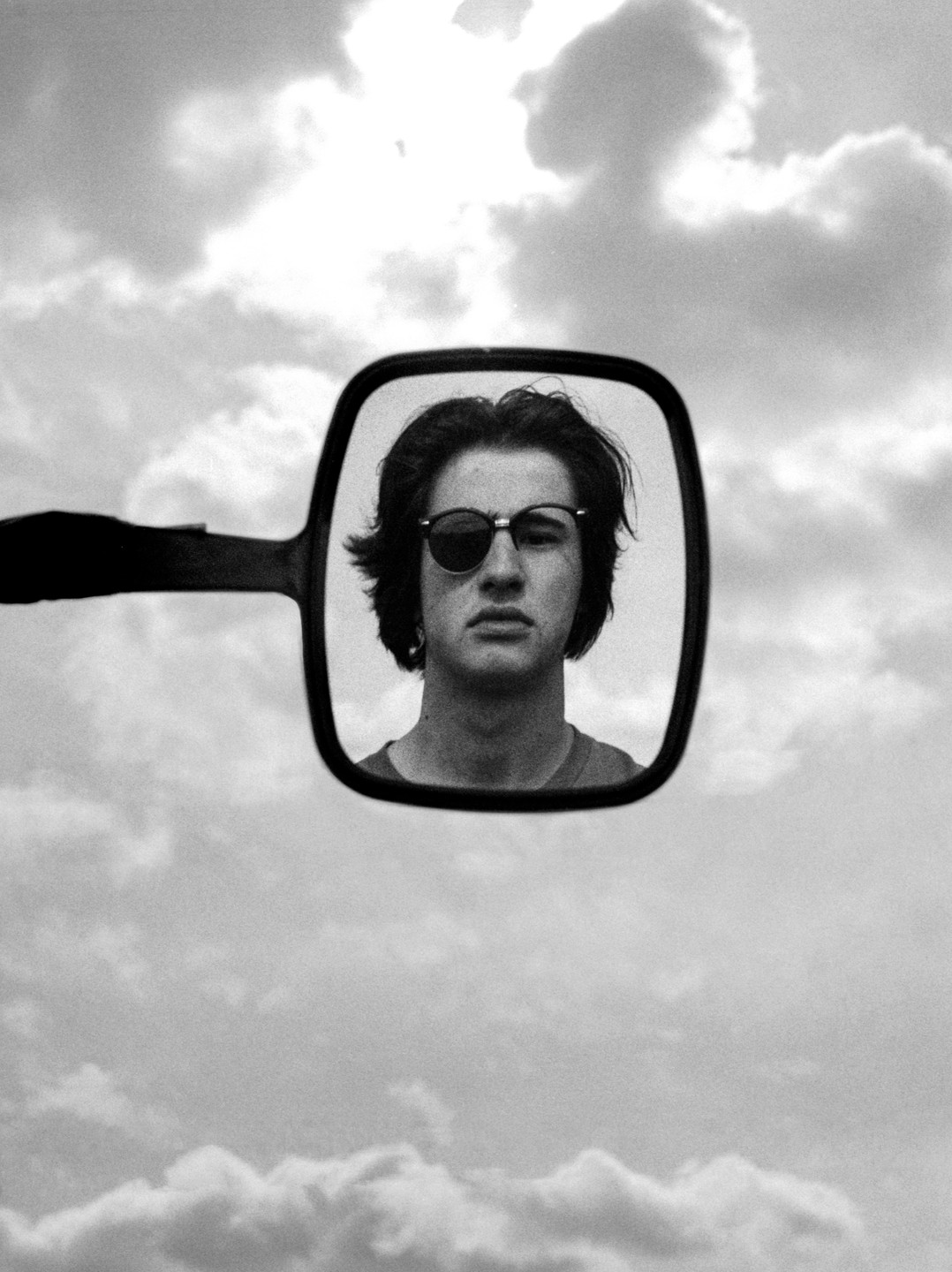 A man wearing sunglasses is reflected in a mirror against a cloudy sky.