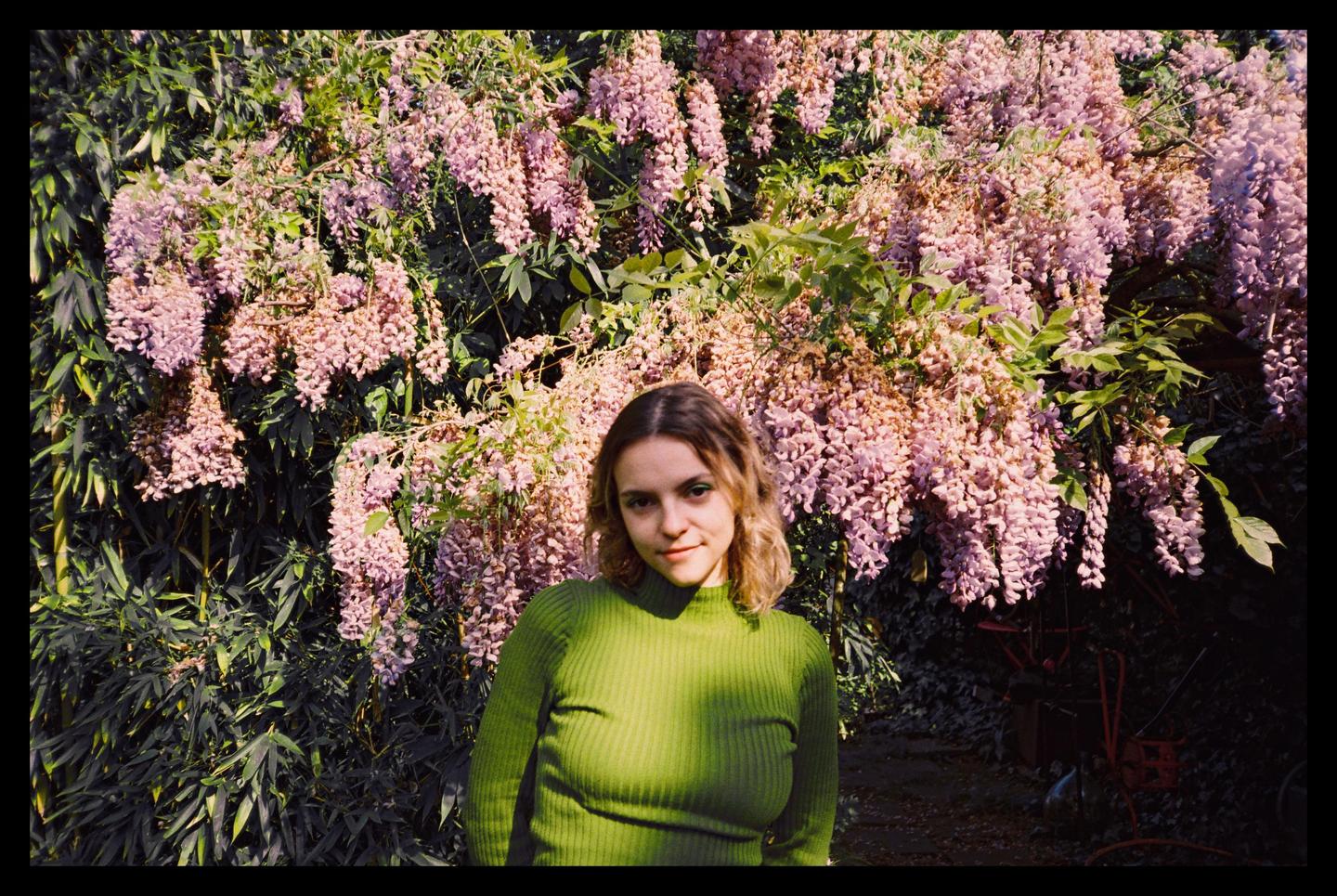 A woman in a green sweater stands in front of a wall of wisteria blossoms.