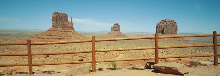 A wooden fence stands in the foreground of a vast desert landscape with three large rock formations under a clear sky.