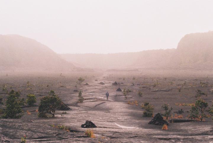 A person stands on a vast, rocky plain with sparse vegetation, surrounded by hazy mountains.