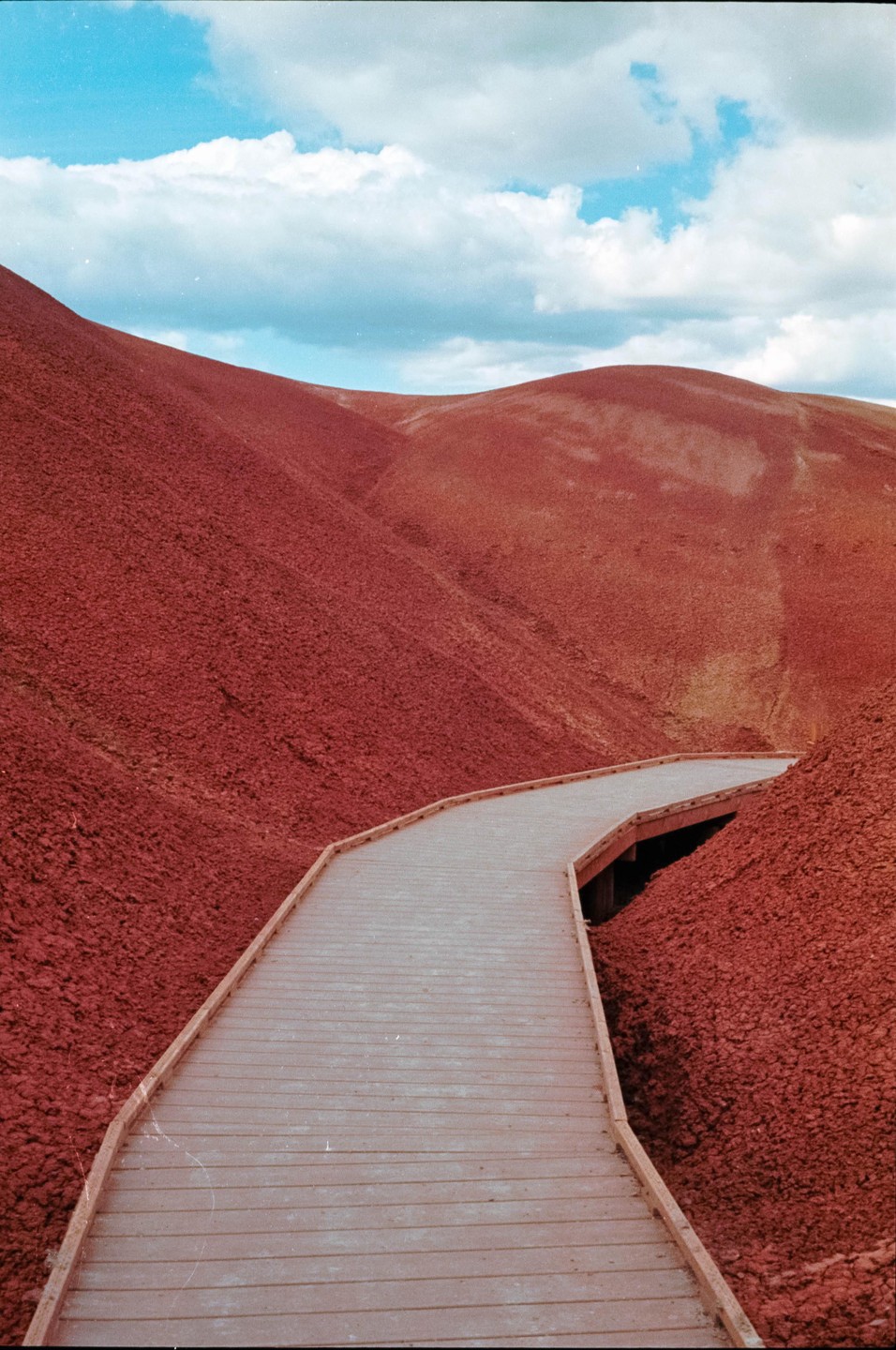 A wooden boardwalk winds through red hills under a blue sky with white clouds.