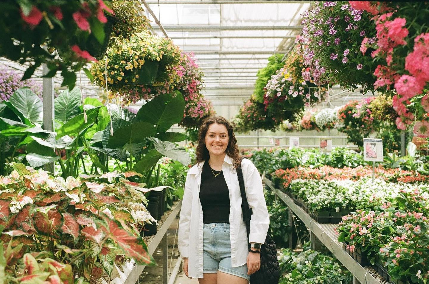 A woman stands smiling in a greenhouse filled with plants and hanging baskets of flowers.