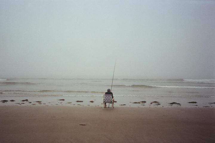 A person sits on a chair on a beach, fishing in the ocean on a hazy day.