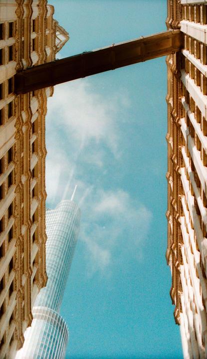 Two ornate buildings frame a modern skyscraper and a skybridge against a bright blue sky.