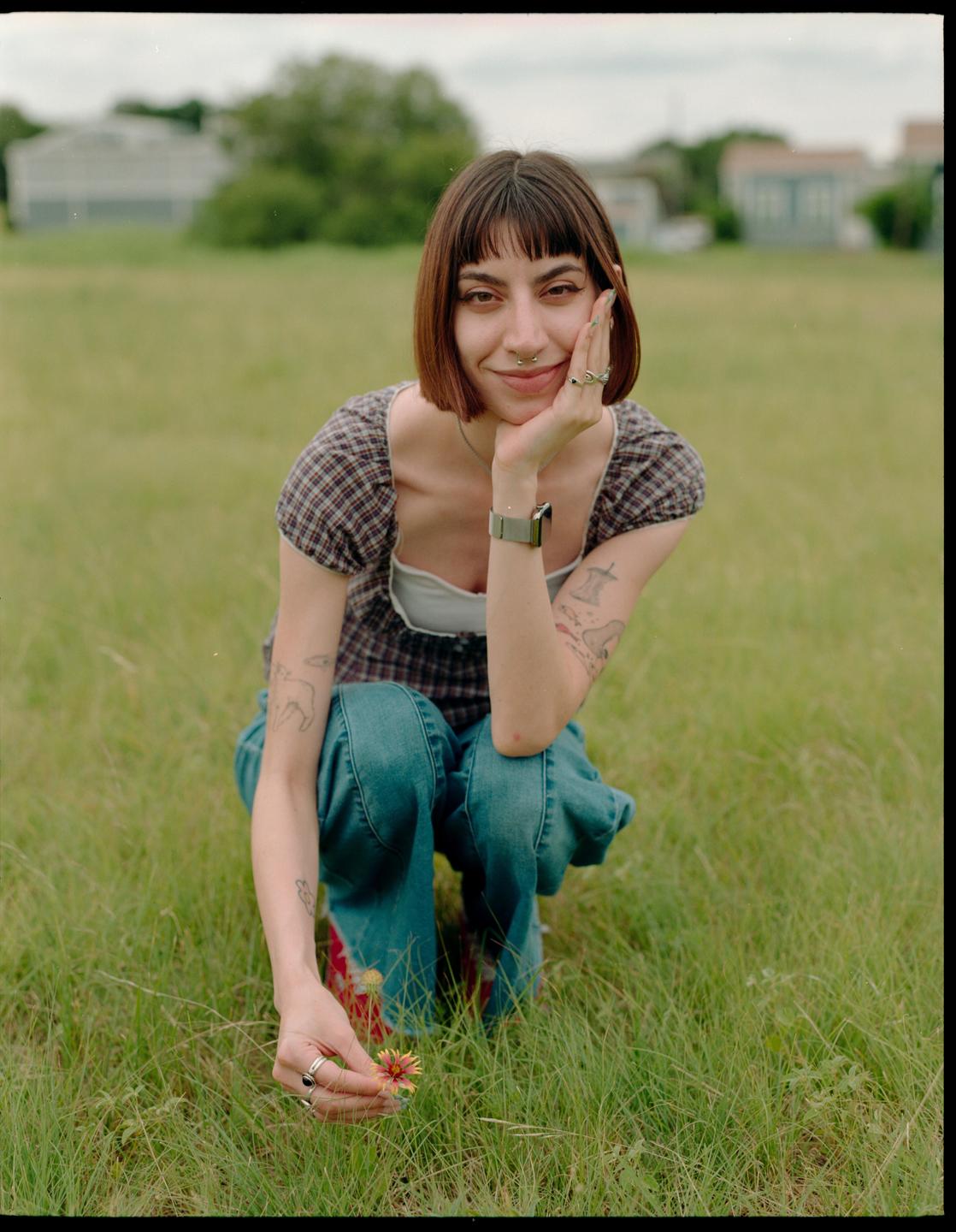 A woman crouches in a grassy field, holding a small flower and looking at the viewer.