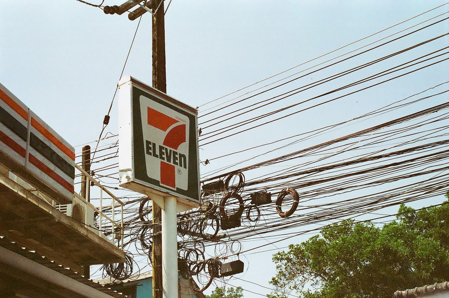 A 7-Eleven sign stands next to a utility pole with many wires against a clear sky.
