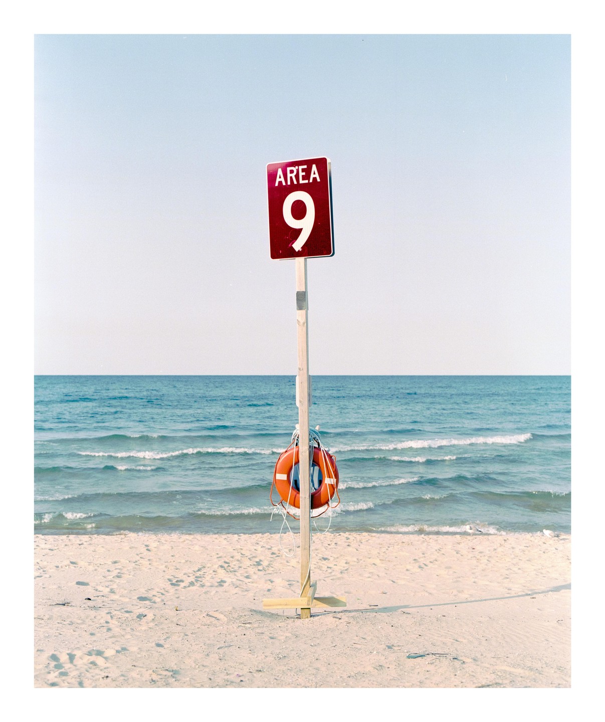 A red sign with "AREA 9" and a life preserver are attached to a wooden post on a sandy beach with the ocean in the background.