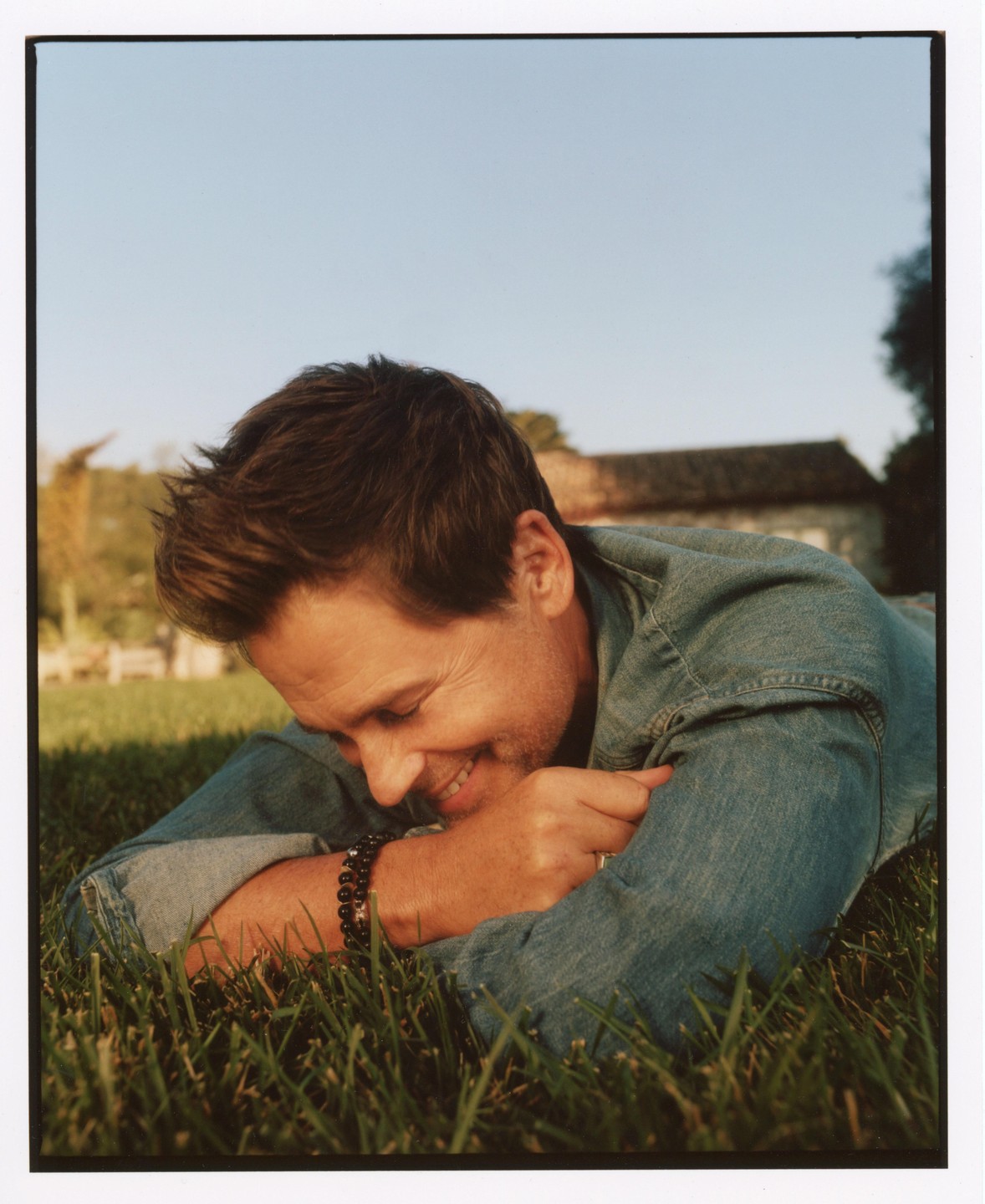 A man in a denim jacket is lying on green grass, smiling with his head resting on his arms.