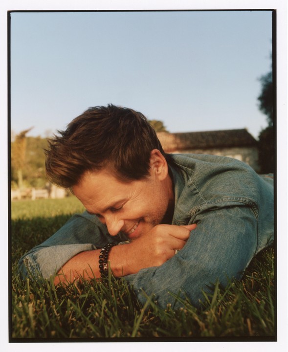 A man in a denim jacket is lying on green grass, smiling with his head resting on his arms.