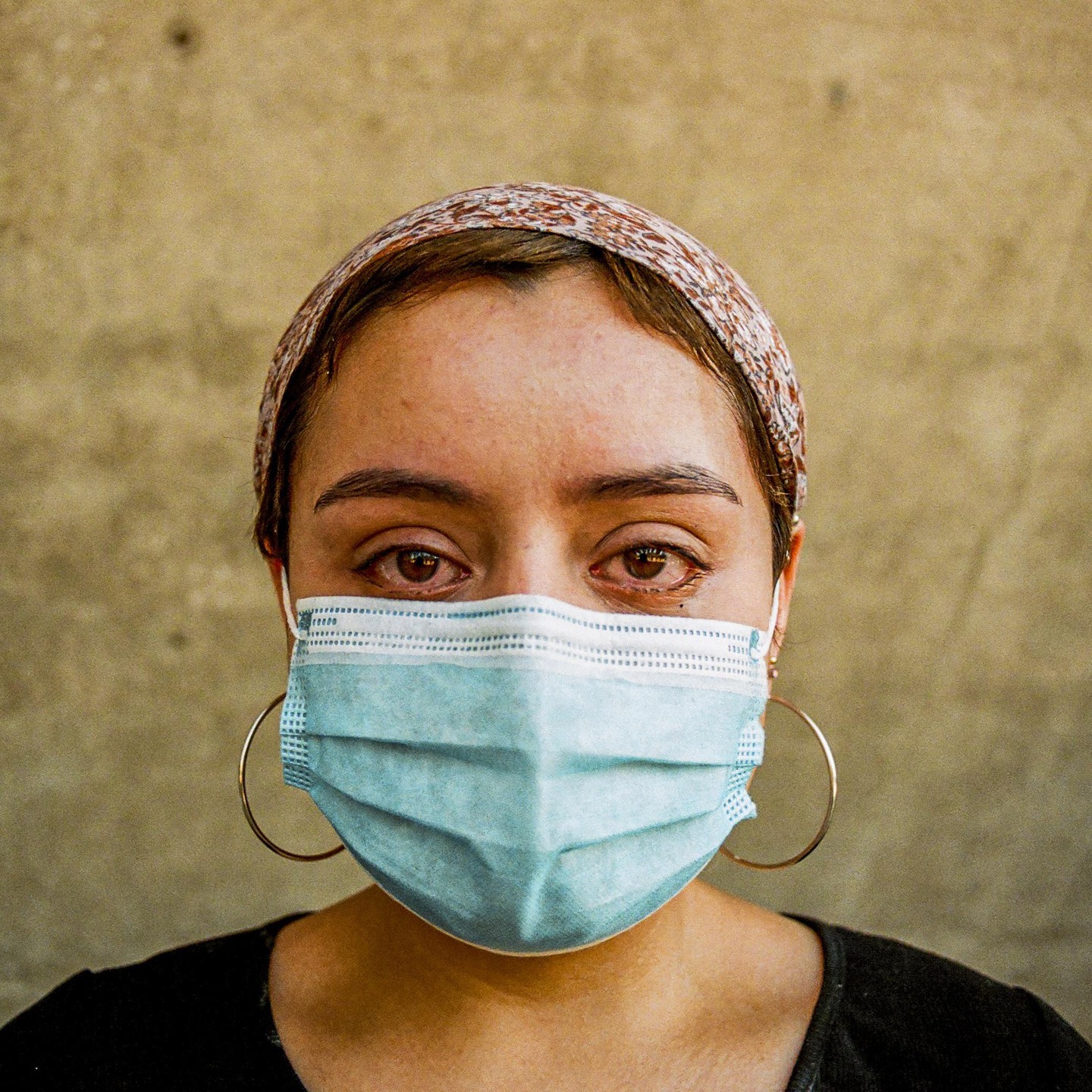 A woman with a patterned headband and large hoop earrings wears a surgical mask and has tears in her eyes.